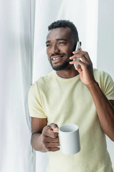 Allegro uomo afroamericano che tiene la tazza con il tè e parla sul cellulare — Foto stock