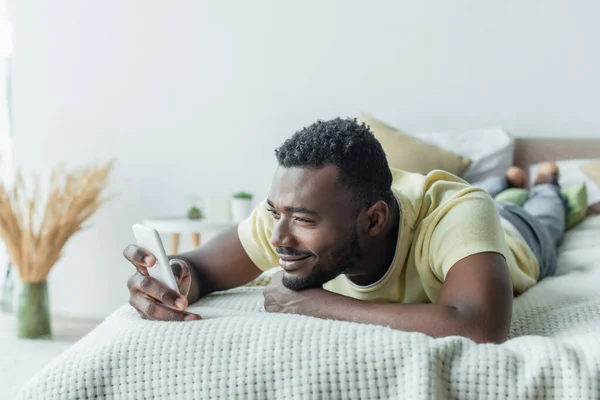 Feliz homem americano africano em t-shirt usando smartphone e deitado na cama — Fotografia de Stock