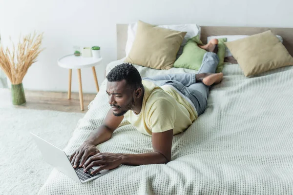 African american freelancer in t-shirt using laptop and lying on bed — Stock Photo