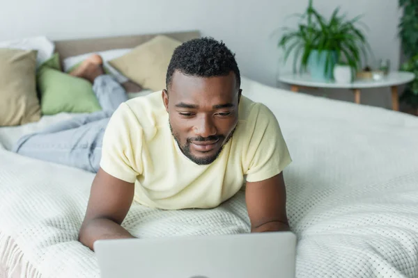 Pleased african american freelancer in t-shirt using laptop and lying on bed — Stock Photo