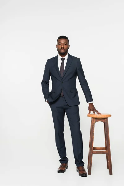 Full length of african american businessman in suit posing with hand in pocket near stool isolated on grey — Stock Photo