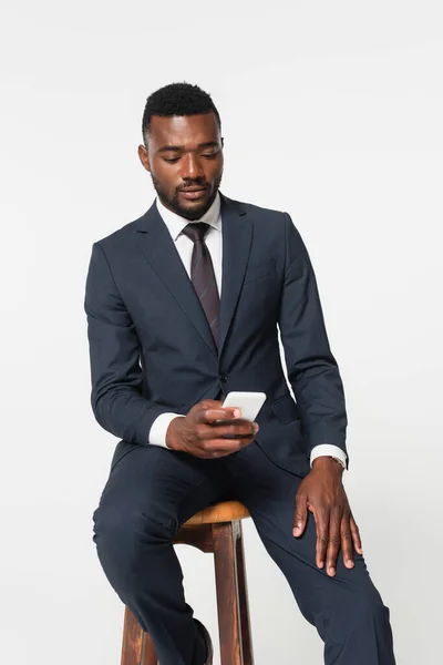 Hombre de negocios afroamericano en traje sentado en taburete de madera y el uso de teléfono inteligente aislado en gris - foto de stock