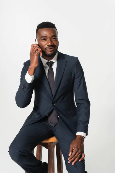 Hombre de negocios afroamericano feliz en traje sentado en taburete de madera y hablando en el teléfono inteligente aislado en gris - foto de stock