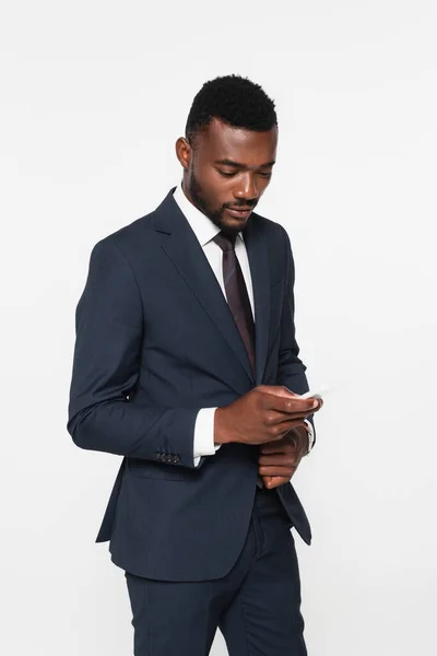 Hombre de negocios afroamericano barbudo en traje de mensajería en el teléfono inteligente aislado en gris - foto de stock