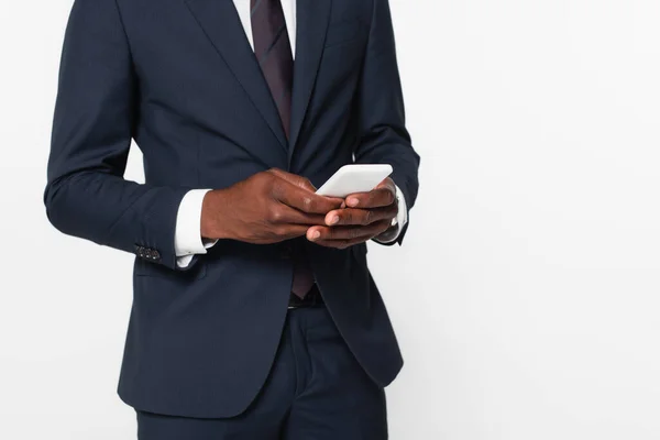 Vista recortada del hombre de negocios afroamericano en traje de mensajería en el teléfono inteligente aislado en gris - foto de stock