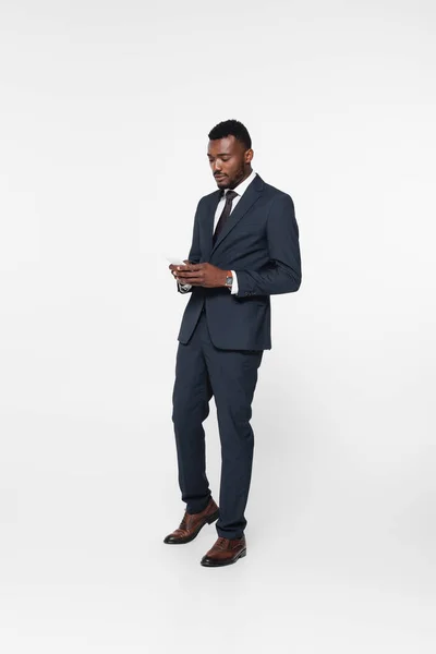 Longitud completa de barbudo hombre de negocios afroamericano en traje mensajes de texto en el teléfono inteligente aislado en gris - foto de stock