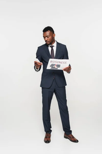 Full length of african american businessman in suit using smartphone while holding newspaper isolated on grey — Stock Photo