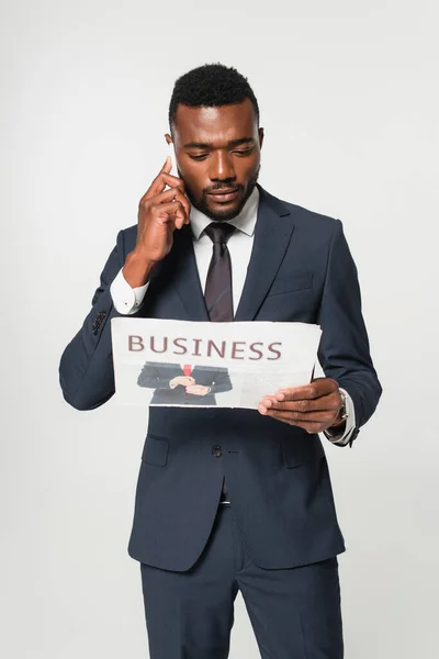 African american man in suit talking on smartphone while reading business newspaper isolated on grey — Stock Photo