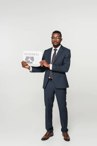 Longitud completa de hombre afroamericano en gafas que muestran periódico de negocios aislado en gris - foto de stock