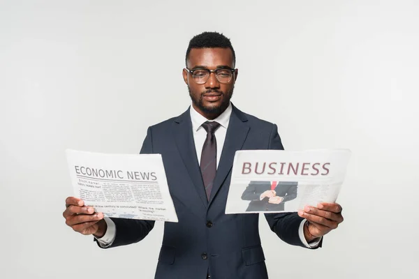 Homem americano africano em óculos segurando jornais com letras de notícias comerciais e econômicas isoladas em cinza — Fotografia de Stock