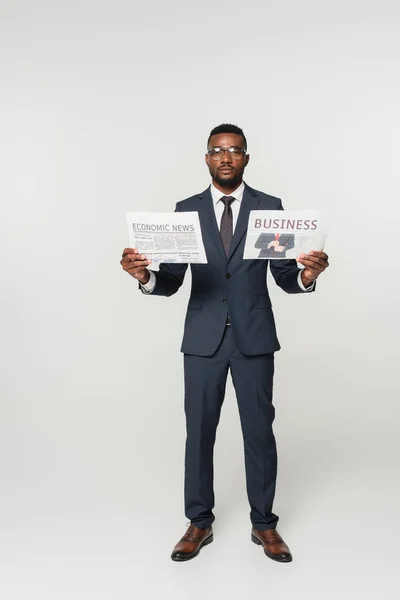 Longitud completa del hombre afroamericano en gafas con periódicos con letras de noticias empresariales y económicas aisladas en gris - foto de stock