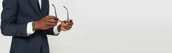 Cropped view of african american man in suit holding eyeglasses isolated on grey, banner — Stock Photo