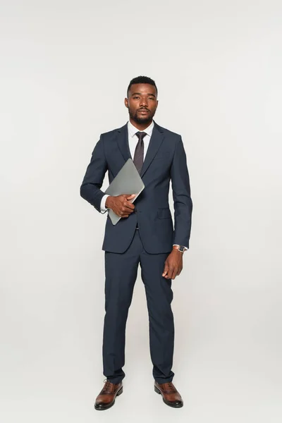 Longitud completa de hombre de negocios afroamericano en traje que sostiene el ordenador portátil aislado en gris - foto de stock