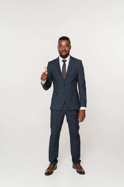 Full length of smiling african american businessman in suit showing thumb up isolated on grey — Stock Photo