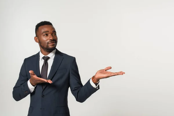 Sonriente hombre de negocios afroamericano en traje apuntando con las manos aisladas en gris - foto de stock