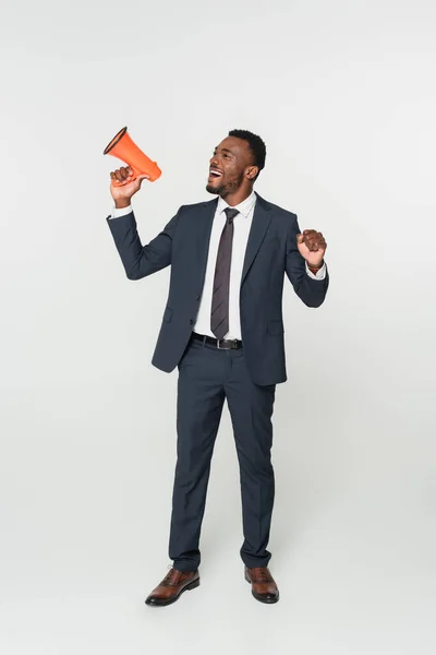 Full length of excited african american businessman in suit screaming in megaphone isolated on grey — Stock Photo