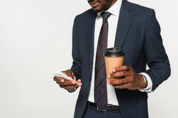 Vue recadrée de l'homme d'affaires afro-américain en costume de messagerie sur smartphone et tenant tasse en papier isolé sur gris — Photo de stock