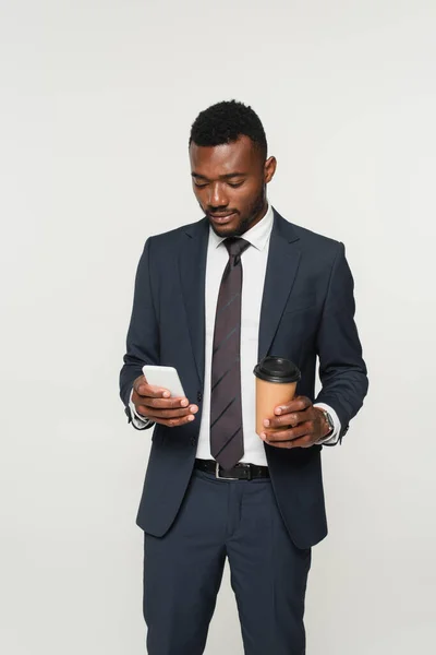 African american businessman in suit messaging on smartphone and holding paper cup isolated on grey — Stock Photo