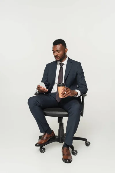 Hombre de negocios afroamericano en traje sentado en la silla de la oficina, mensajería en el teléfono inteligente y sosteniendo taza de papel aislado en gris - foto de stock