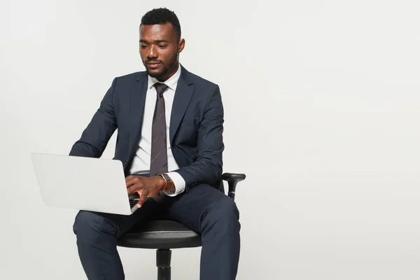 Афріканський американський бізнесмен в костюмі сидячи в офісному кріслі і використовуючи ноутбук ізольований на сірому — стокове фото