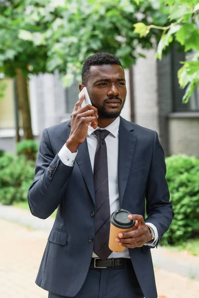 Bearded african american businessman in suit holding paper cup and talking on cellphone — Stock Photo