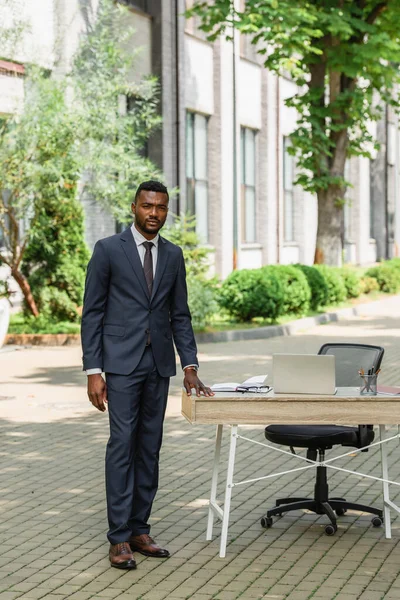 Африканский американский бизнесмен в костюме стоит у стола с ноутбуком на открытом воздухе — стоковое фото
