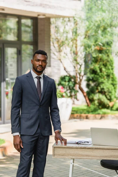 Homme d'affaires afro-américain barbu en costume debout près du bureau avec ordinateur portable à l'extérieur — Photo de stock