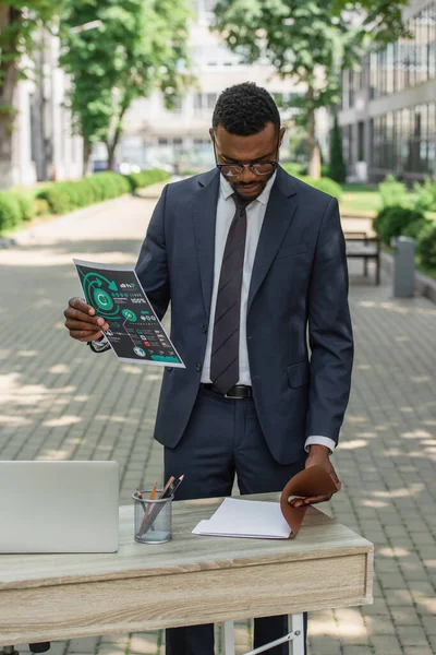 Африканский американский бизнесмен в очках смотрит в папку, держа в руках напечатанную инфографику — стоковое фото