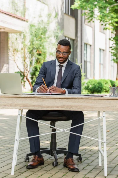 Веселый африканский американский бизнесмен в очках пишет в блокноте возле ноутбука — стоковое фото