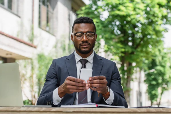 Bearded african american businessman in eyeglasses using smartphone near laptop — Stock Photo