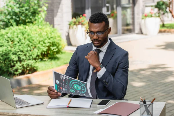 Бородатий афроамериканський бізнесмен в окулярах дивиться на інфографіку біля гаджетів на столі — стокове фото