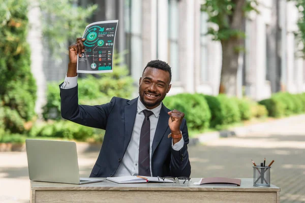 Happy african american businessman holding infographics near laptop on desk — Stock Photo