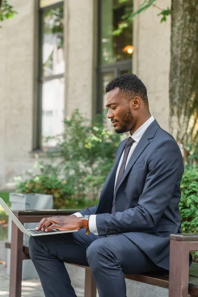 Barbudo hombre de negocios afroamericano escribiendo en el ordenador portátil mientras está sentado en el banco - foto de stock