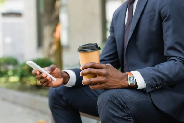 Vista recortada del hombre de negocios afroamericano usando teléfono móvil y sosteniendo café para salir - foto de stock