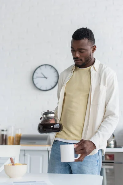 Uomo africano americano che tiene la caffettiera vicino alla tazza in cucina — Foto stock