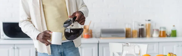 Cropped view of african american man holding coffee pot near cup in kitchen, banner — Stock Photo
