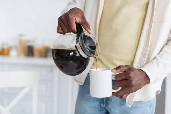 Vue recadrée d'un homme afro-américain tenant une cafetière et versant une boisson dans une tasse — Photo de stock