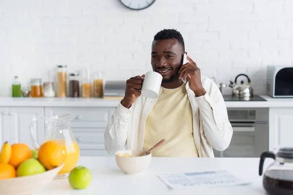 Happy african american man holding cup of coffee and talking on smartphone in kitchen — Stock Photo