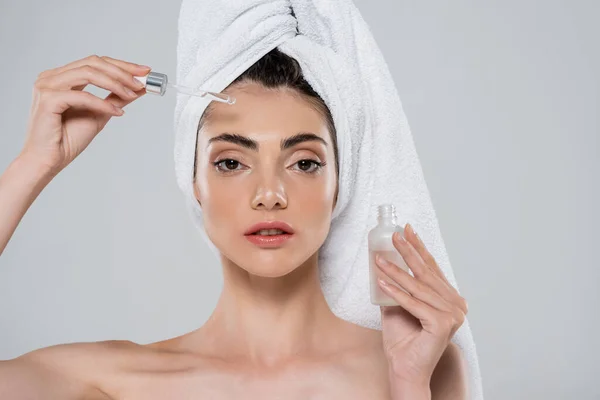 Jeune femme avec serviette sur la tête appliquant sérum hydratant avec pipette isolé sur gris — Photo de stock