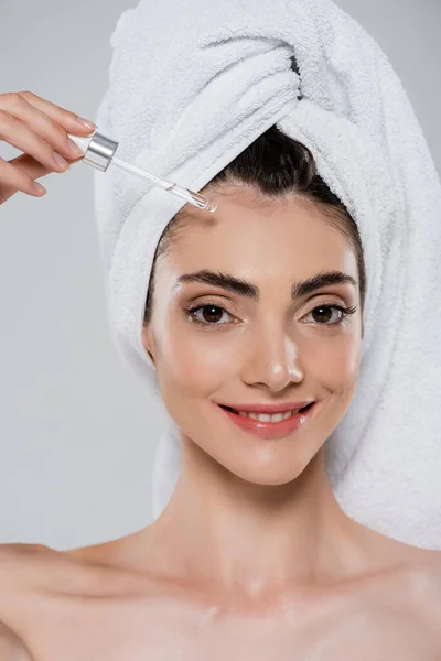 Sonriente joven con toalla en la cabeza aplicando suero hidratante con pipeta aislada en gris - foto de stock