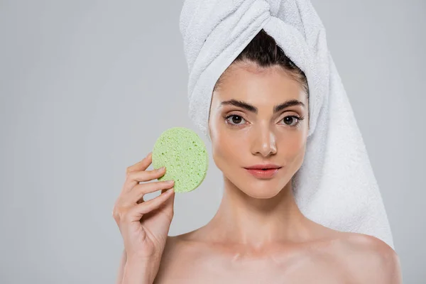 Junge Frau mit Handtuch auf dem Kopf hält grünen Schönheitsschwamm isoliert auf grau — Stockfoto