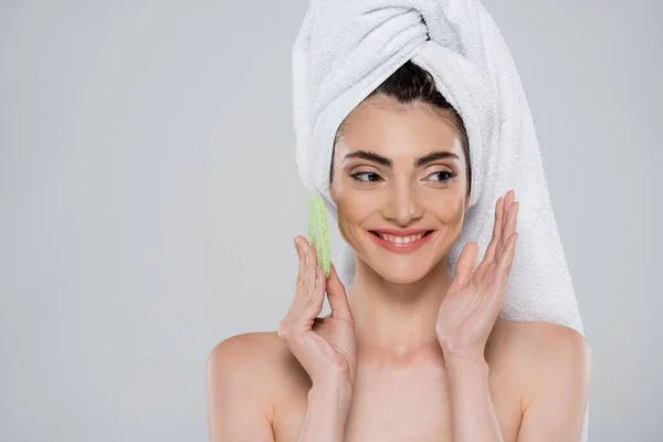 Cheerful young woman with towel on head holding green beauty sponge isolated on grey — Stock Photo