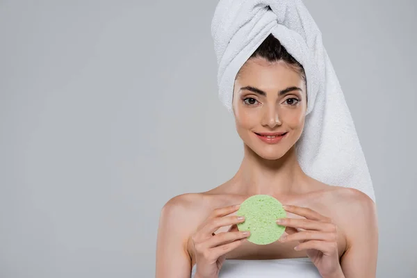 Улыбающаяся молодая женщина с полотенцем на голове держа зеленую губку красоты изолированы на сером — стоковое фото