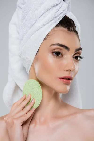 Pretty young woman with towel on head holding green beauty sponge isolated on grey — Stock Photo