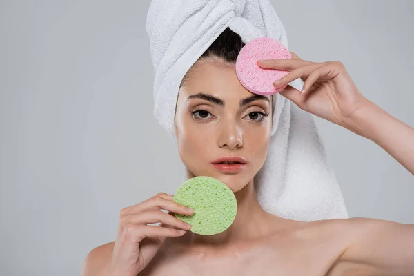 Young woman with towel on head holding beauty sponges isolated on grey — Stock Photo