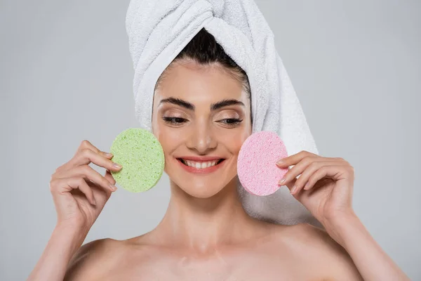 Heureuse jeune femme avec serviette sur la tête tenant des éponges de beauté isolé sur gris — Photo de stock