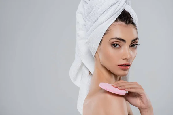 Jeune femme avec serviette sur la tête tenant l'éponge de beauté rose et regardant la caméra isolée sur gris — Photo de stock