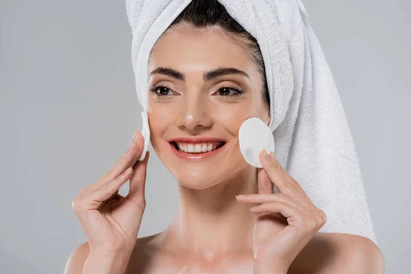 Счастливая женщина с полотенцем на голове удаление макияжа с хлопчатобумажными подушечками изолированы на сером — стоковое фото