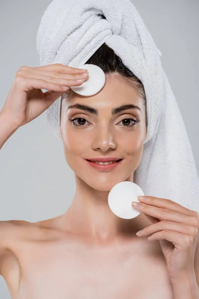 Mulher sorridente com toalha na cabeça removendo maquiagem com almofadas de algodão isoladas em cinza — Fotografia de Stock