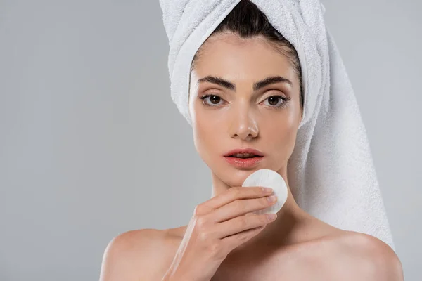 Mujer joven con toalla en la cabeza quitando maquillaje con almohadilla de algodón aislada en gris - foto de stock